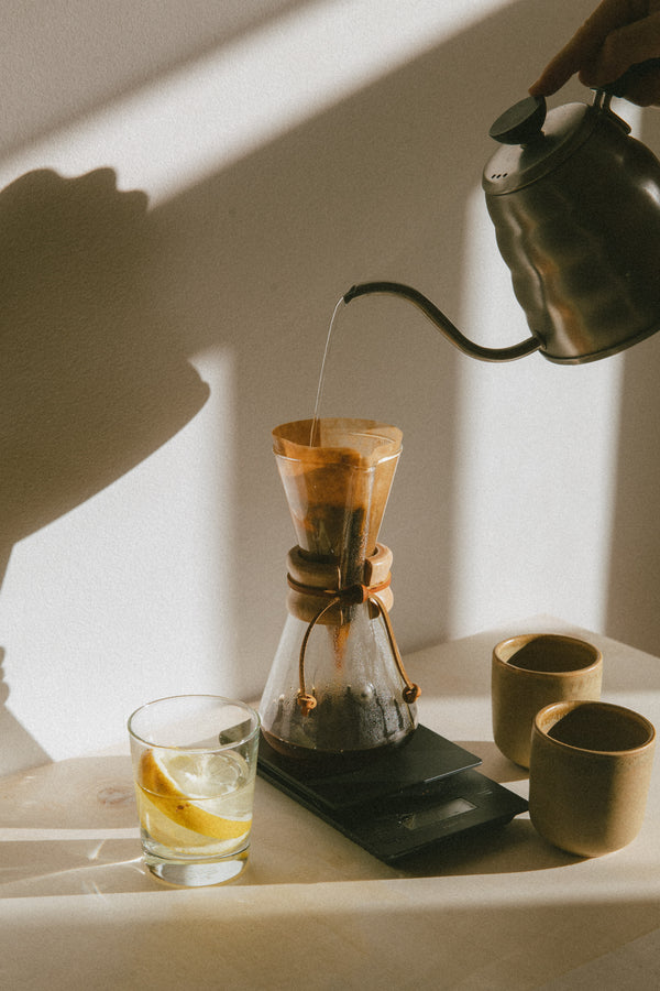 Person pouring liquid from a teapot into a glass coffee maker on a table with a glass of lemon water.